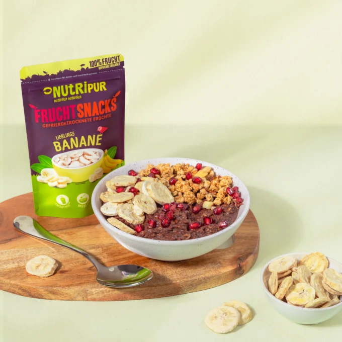 Hier ist ein Bild mit gefriergetrockneten Bananen als Müsli-Topping und Porridge-Topping in einer Schale. Im Hintergrund ist die Verpackung abgebildet.