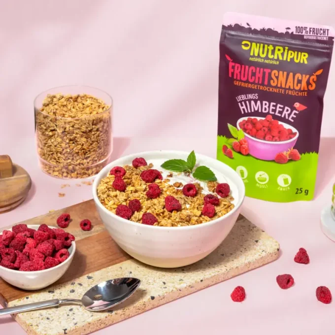 Hier ist ein Bild mit gefriergetrockneten Himbeeren mit Müsli und Joghurt in einer Müslischale. Im Hintergrund ist die Verpackung abgebildet.