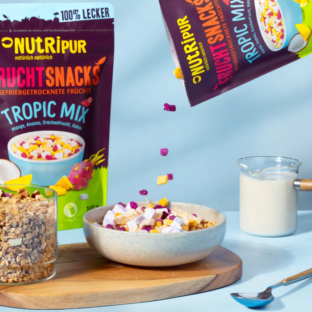 Hier ist ein Bild mit gefriergetrocknetem Tropic-Fruchtmix in einer Schale mit Joghurt. Im Hintergrund ist die Verpackung abgebildet.