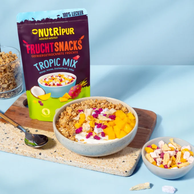 Hier ist ein Bild mit gefriergetrockneten Früchten des Nutripur Tropic-Mix als Müsli-Topping. Im Hintergrund ist die Verpackung abgebildet.